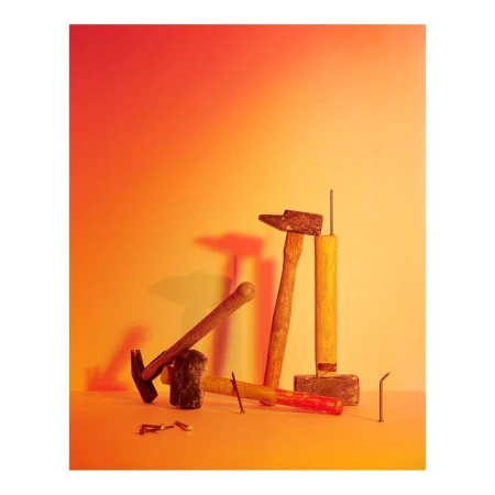 Still life composition featuring hammers, nails, and other tools against a gradient orange and yellow background in a studio setting.