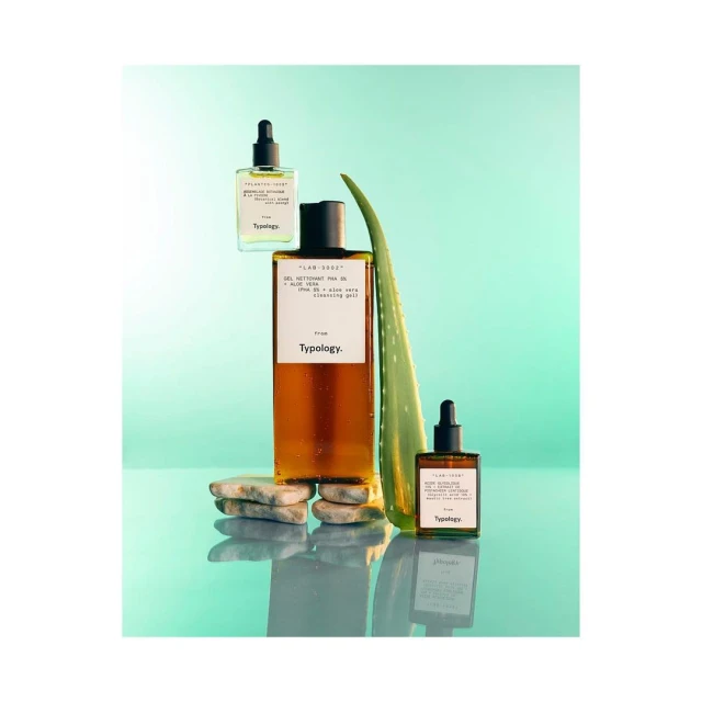 Still life shot of Typology skincare products, including serums and cleansing gel, arranged with aloe vera leaf on a reflective surface.