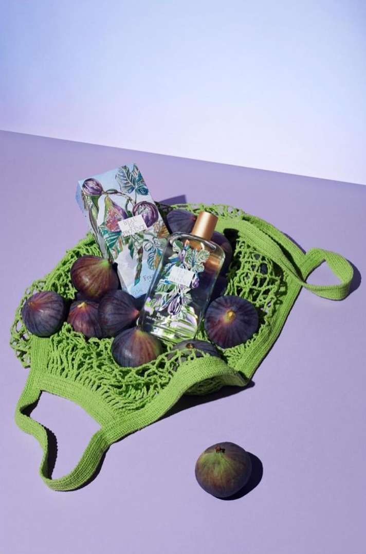 Still life of perfume bottle, patterned box, and figs in a green woven bag on a lilac surface with a gradient white to lilac background.
