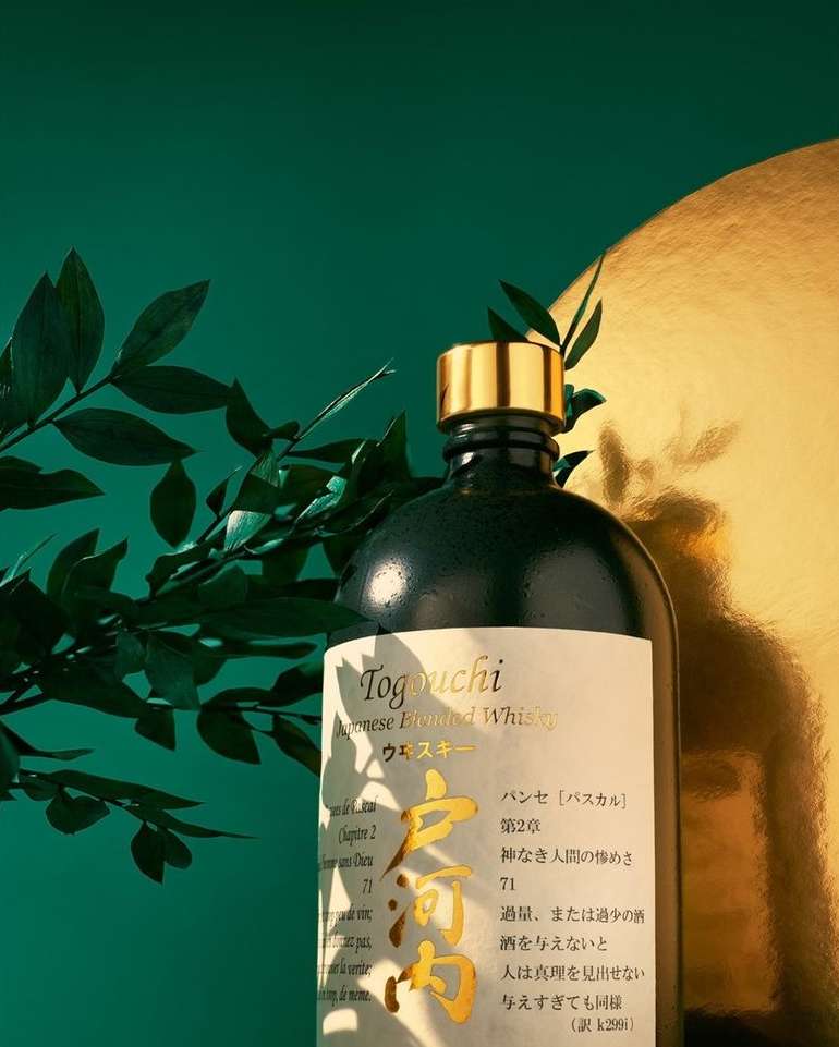 Studio shot of Togouchi Japanese Blended Whiskey bottle with gold cap and label, green leaves, and backdrop with gold accent.
