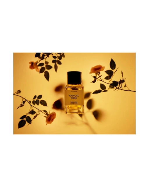 Studio shot of a perfume bottle with RADICAL ROSE printed on the label surrounded by rose branches with blossoms against a yellow background.