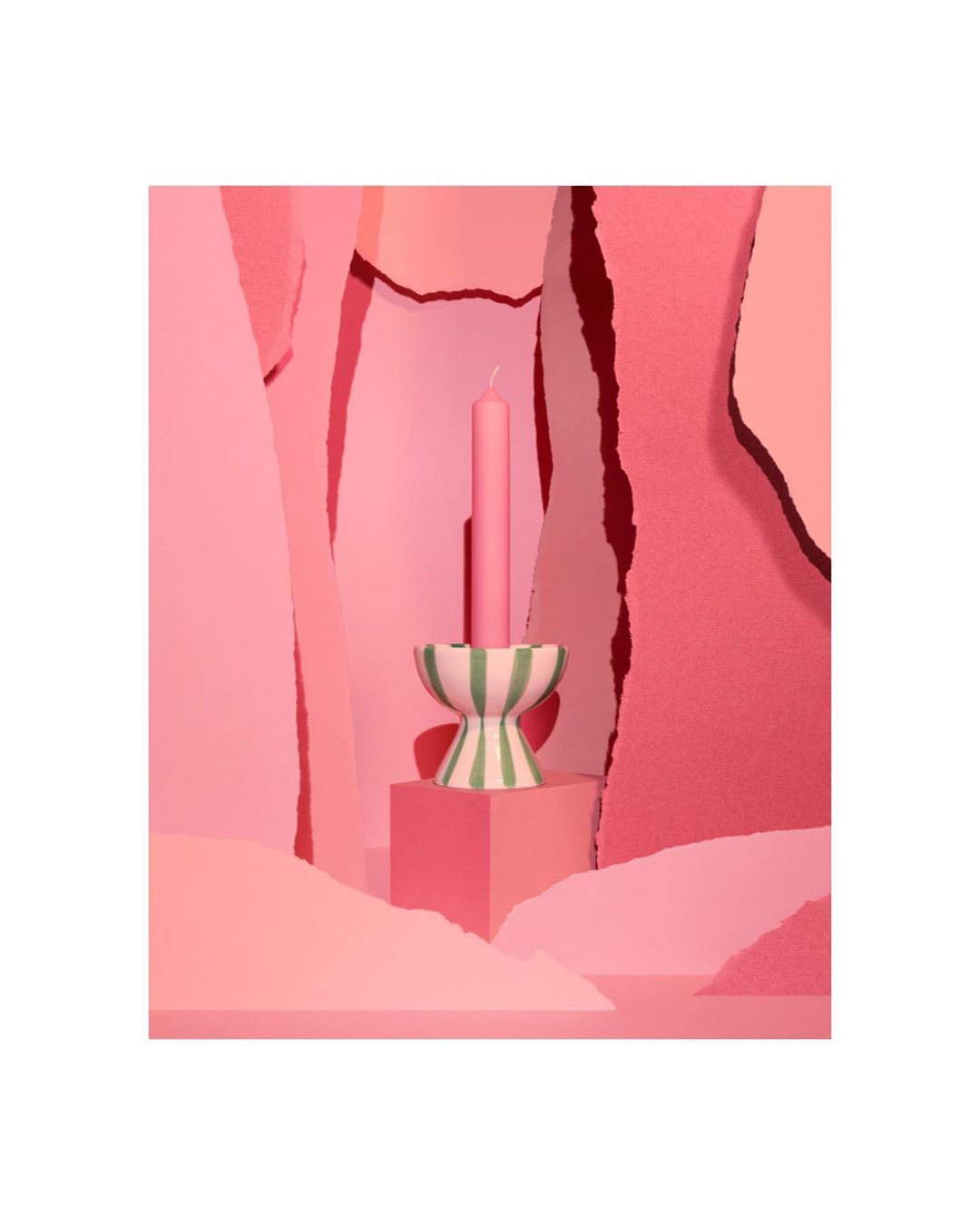 Pink candle in a striped candle holder sits on a cube against a geometric background in various shades of pink for a playful, modern look.