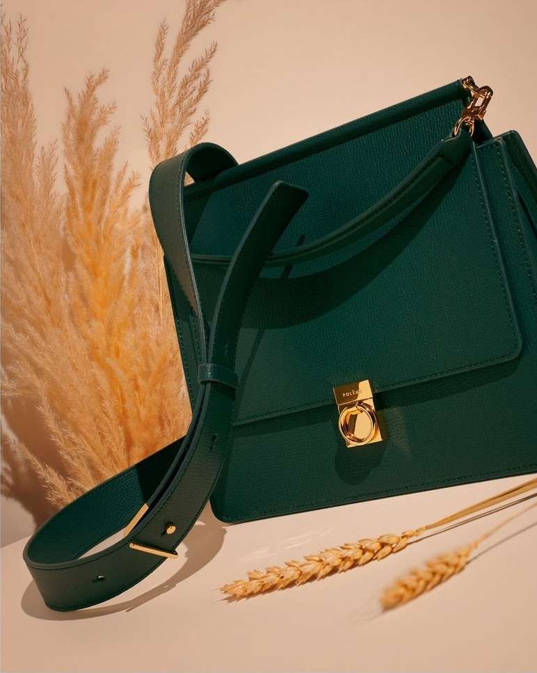 Elegant green leather satchel-style handbag with gold hardware and shoulder strap, styled with wheat stalks against a light beige backdrop.