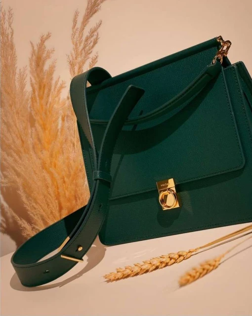 Elegant green leather satchel-style handbag with gold hardware and shoulder strap, styled with wheat stalks against a light beige backdrop.