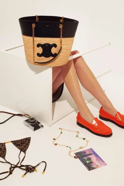 Straw tote bag with black leather trim sits on a white cube near bikini top, necklace, postcard, and red loafers.
