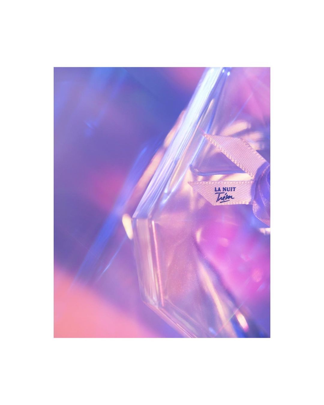 Close-up of a perfume bottle with a white ribbon and the brand name, showcasing a blend of pink and blue hues, reflecting modern beauty trends.
