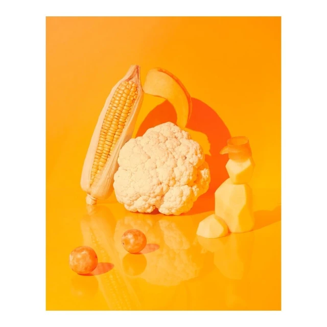 Still life of corn on the cob, cauliflower, plums, and potato pieces on a reflective orange surface, brightly lit with a strong shadow.