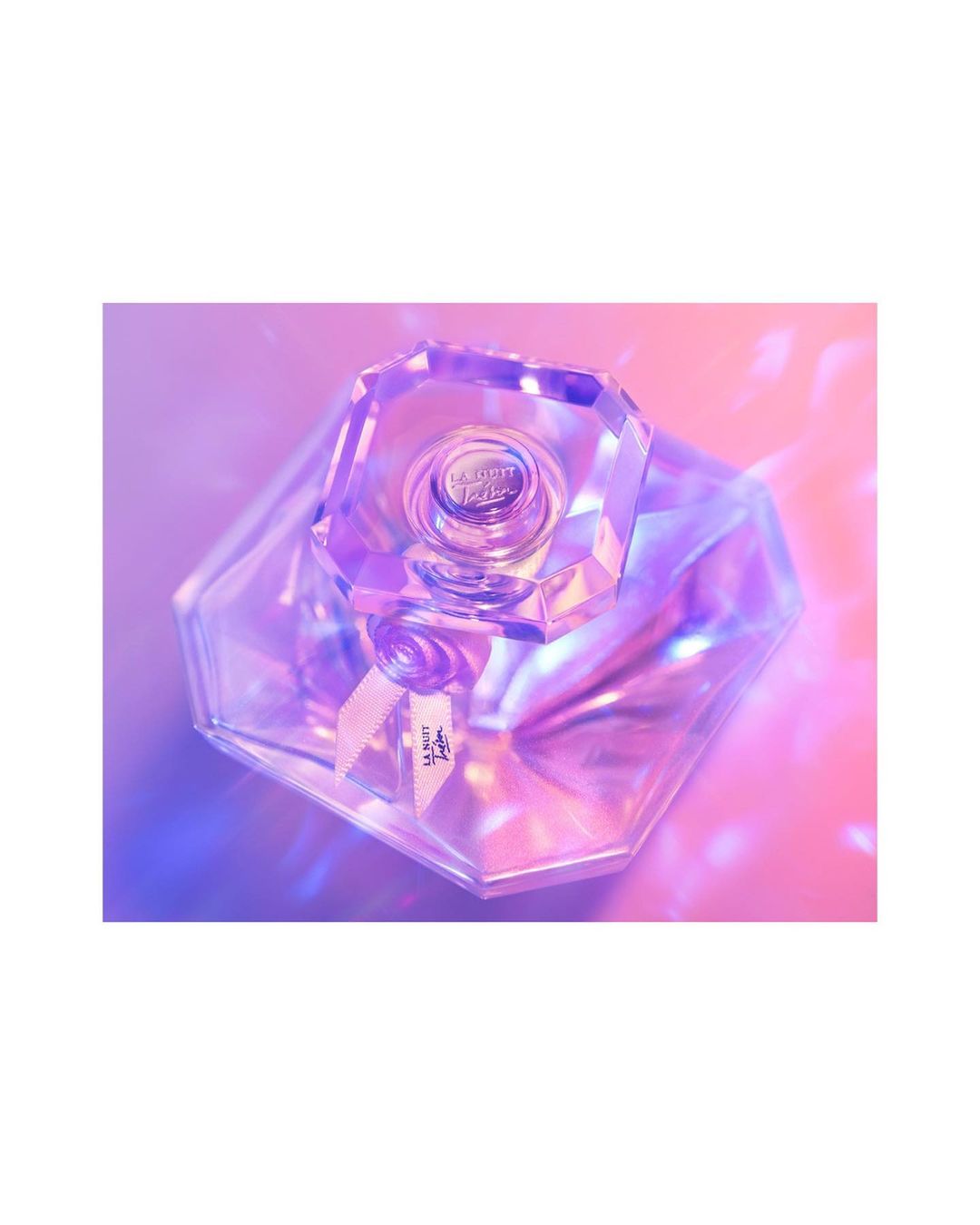 Close-up product shot of a crystal perfume bottle on a pink and purple gradient background with geometric light patterns. Beauty product.