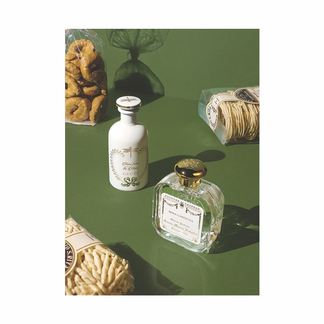 Two Gucci perfumes stand on a green surface alongside pasta and crackers. The perfumes are in white and transparent bottles with gold accents.