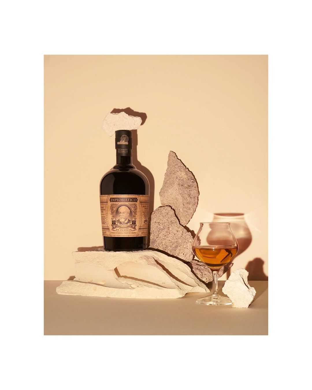 A still-life photograph featuring a bottle of Diplomático rum, a tasting glass with amber liquid, and decorative stone pieces against a neutral backdrop.