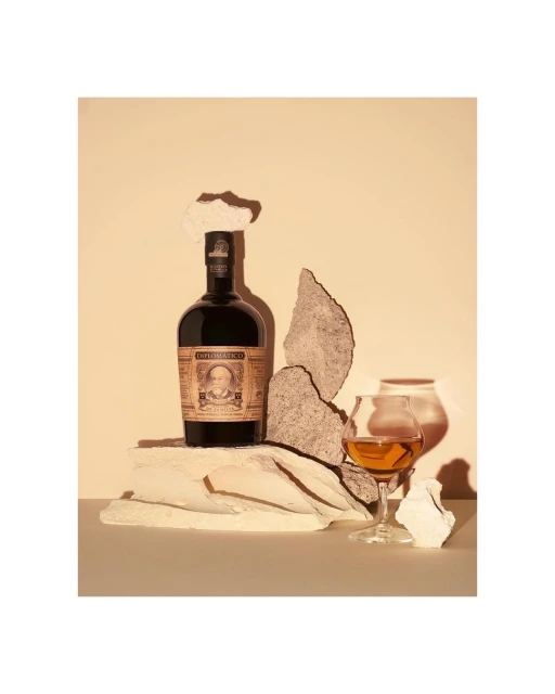 A still-life photograph featuring a bottle of Diplomático rum, a tasting glass with amber liquid, and decorative stone pieces against a neutral backdrop.