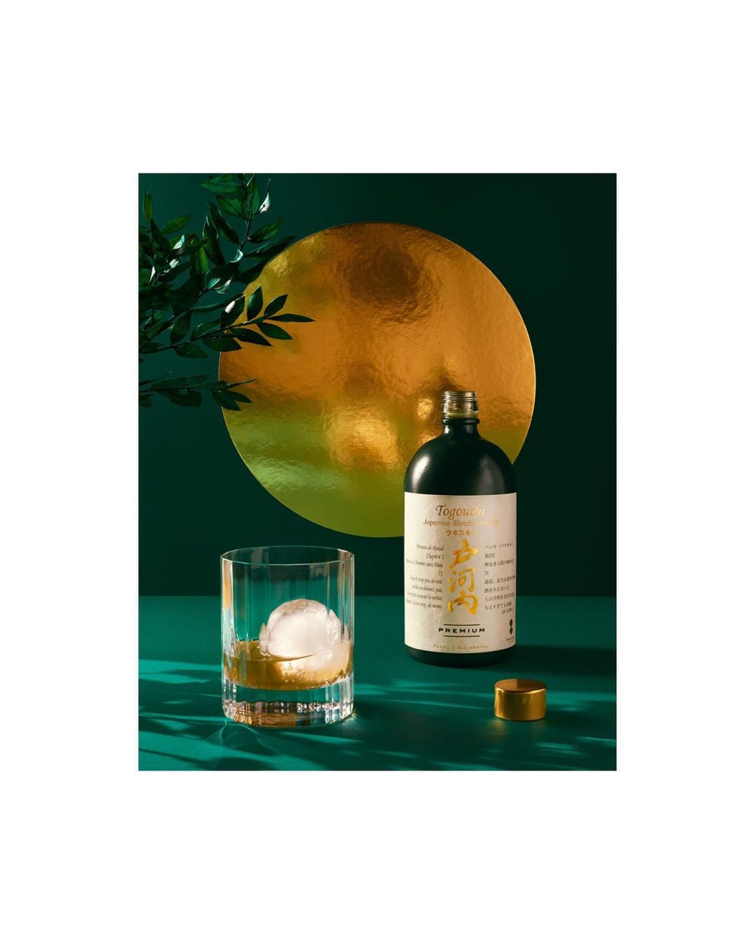 Crystal glass of amber liquid with ice sphere and a bottle set against a dark green backdrop and a golden disc.