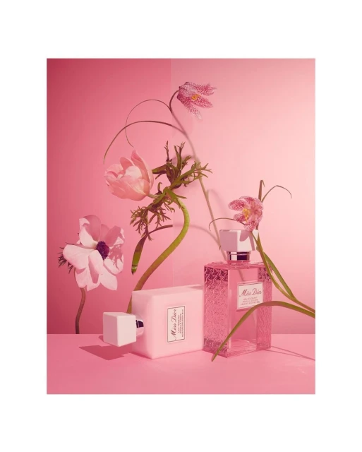 Two Miss Dior perfume bottles, one rectangular and one cube-shaped, are surrounded by flowers and greenery against a pink background.