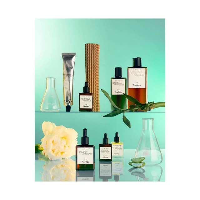 Still life shot of Typology skincare products on glass shelving against a teal background, featuring dropper bottles, cream, bamboo stalks, and laboratory beakers.