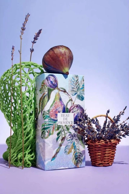 Artistic still life composition featuring a fig fruit atop a Beau de Provence box, complemented by lavender and a green mesh bag.