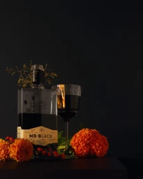 Bottle of Mr. Black coffee liqueur and a wine glass filled with dark liquid surrounded by orange flowers on a dark surface.