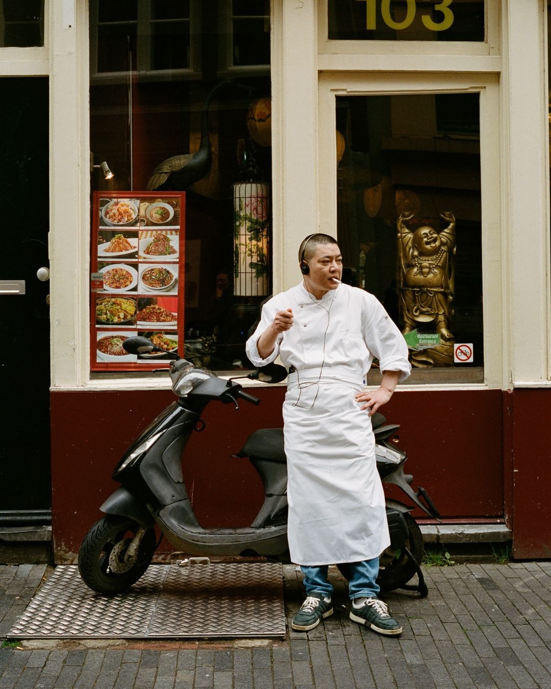 Chef in uniform stands smoking on a street near a black scooter and a restaurant with an image of prepared dishes on display.