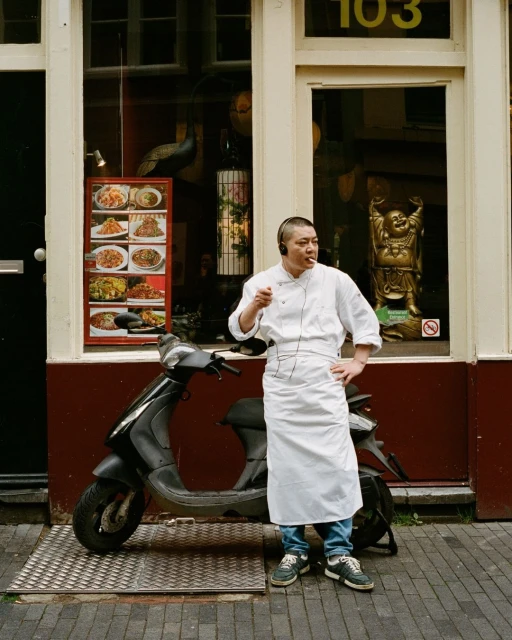 Chef in uniform stands smoking on a street near a black scooter and a restaurant with an image of prepared dishes on display.