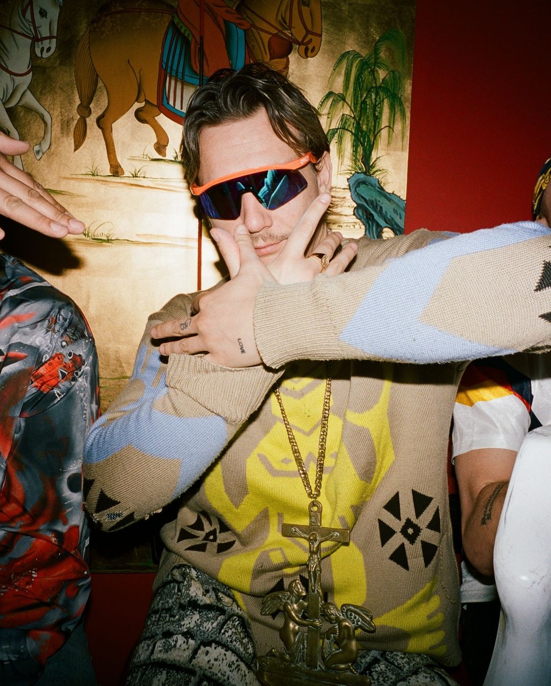 Close-up of a man with sunglasses, a patterned sweater, and a large cross necklace, posing with his hands up, indoors.