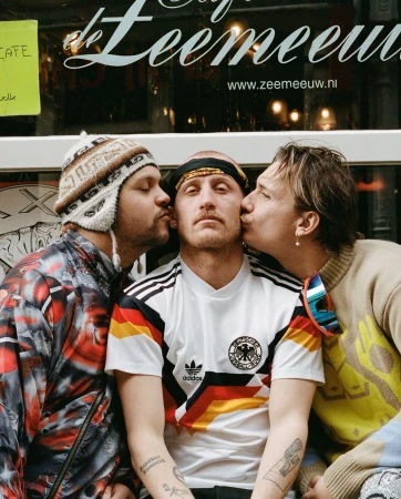 Three young men posing together, with two men kissing the cheeks of a central man wearing a German football jersey in Amsterdam.