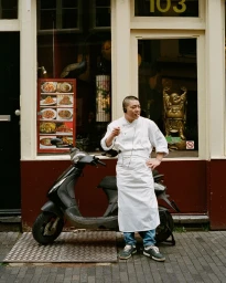 Chef in white jacket and apron standing beside a black scooter outside a restaurant facade in Amsterdam directed by Camille Boumans.