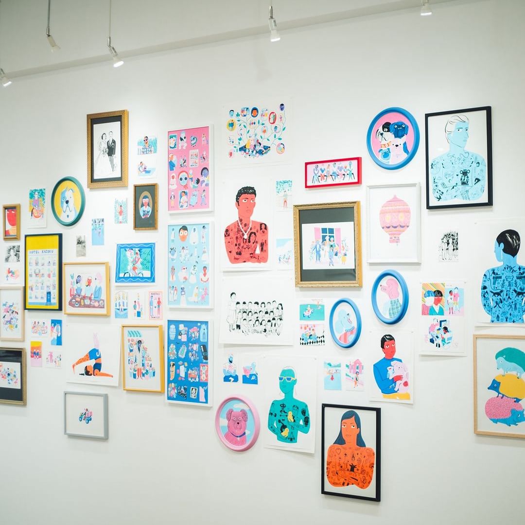 Artwork display featuring a diverse collection of framed illustrations and prints mounted on a white wall in an indoor space.