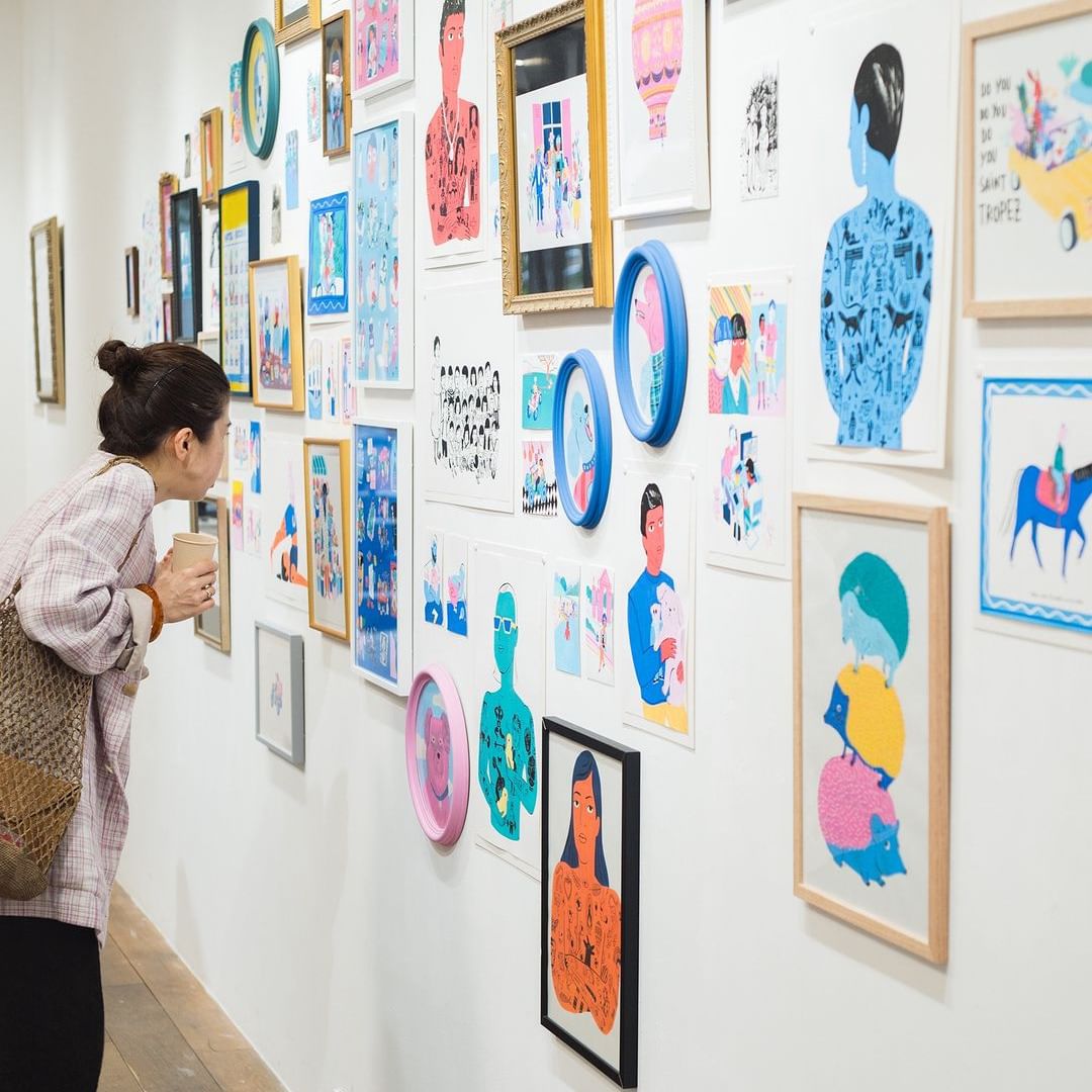 Woman views an art gallery exhibit featuring framed, colorful illustrations and designs hanging on a white wall, creating a vibrant display.