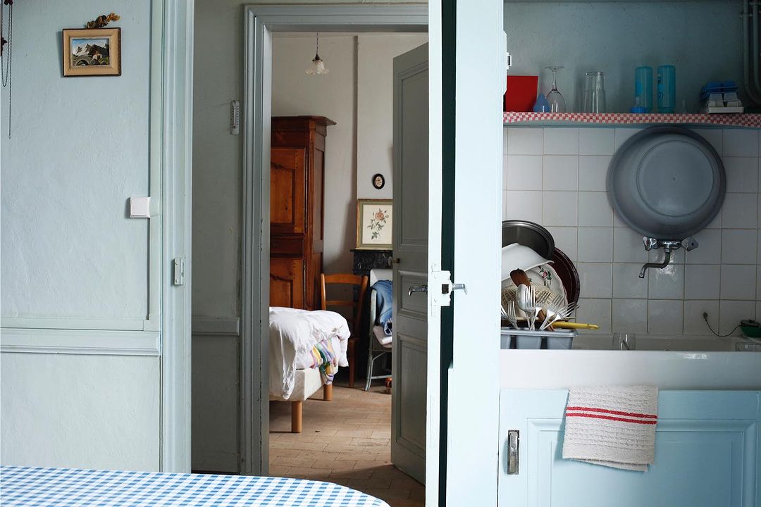 Interior view through a doorway, showing a bedroom in the background and a kitchen sink area in the foreground, bathed in soft light.