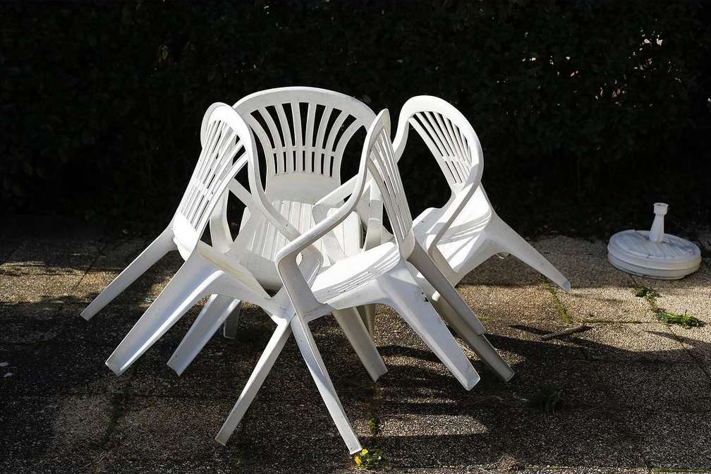 Stacked white plastic chairs sit on a stone patio in sunlight. Background shows foliage and a white patio umbrella base.