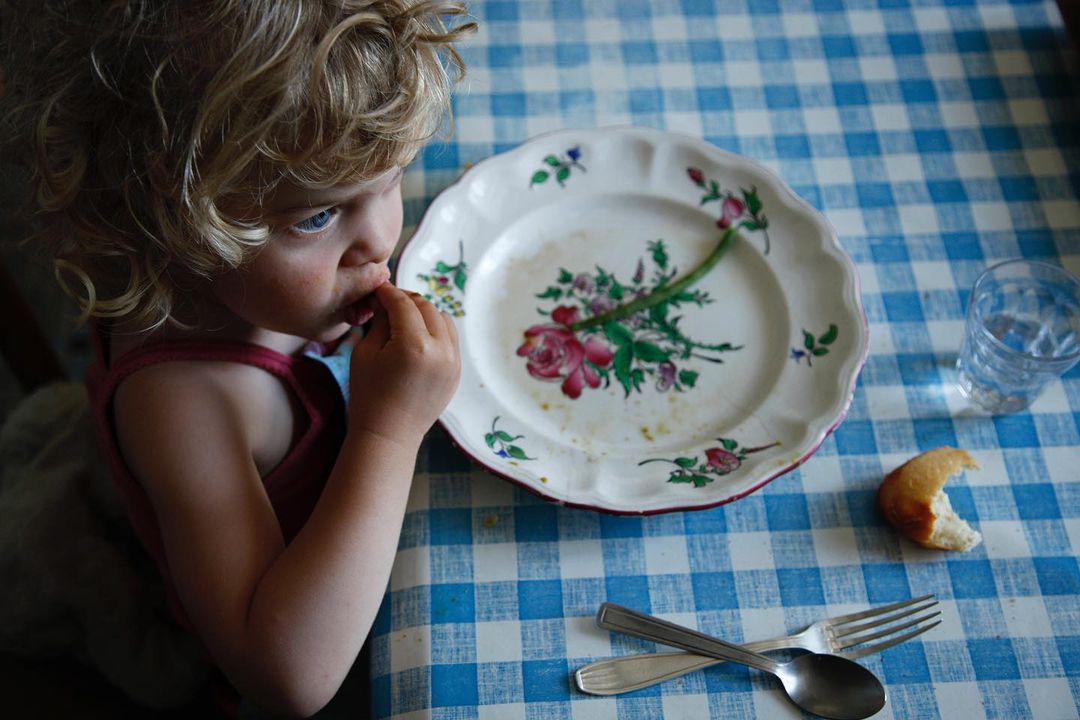 A child with curly blonde hair sits at a blue checkered tablecloth, eating. A plate, glass, cutlery, and a piece of bread are visible.