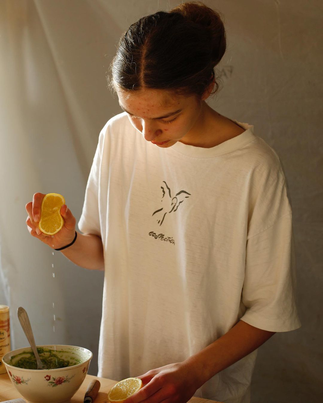 Young adult female squeezes lemon juice into a floral bowl with a spoon and green sauce at a wooden table indoors during daytime.
