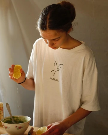 Young adult female squeezes lemon juice into a floral bowl with a spoon and green sauce at a wooden table indoors during daytime.