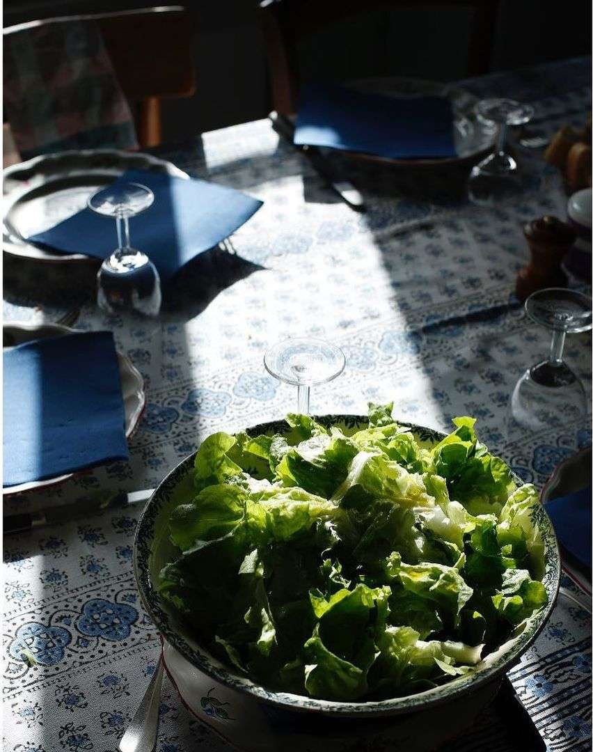 A bowl of fresh lettuce salad sits on a table with blue patterned cloth, complemented by stemware and natural light in an indoor setting.