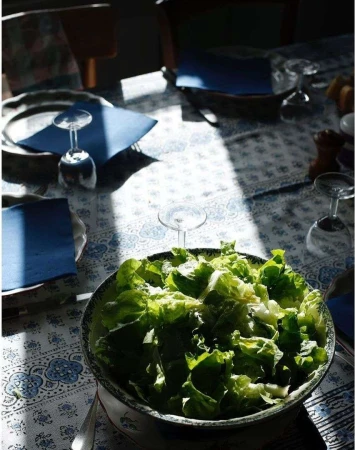 A bowl of fresh lettuce salad sits on a table with blue patterned cloth, complemented by stemware and natural light in an indoor setting.