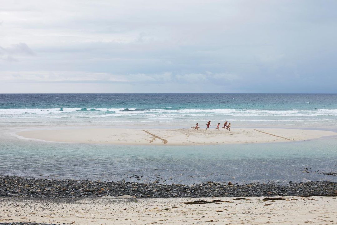 Children run along a sandy beach near the ocean under an overcast sky. Waves break in the distance, and the shoreline is visible.