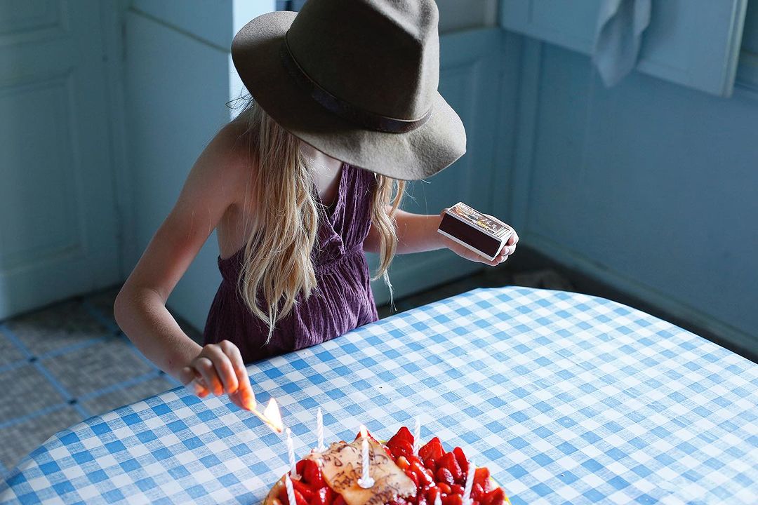 A young girl in a hat lights birthday candles on a strawberry-covered cake with a match, set on a blue-and-white checkered tablecloth.