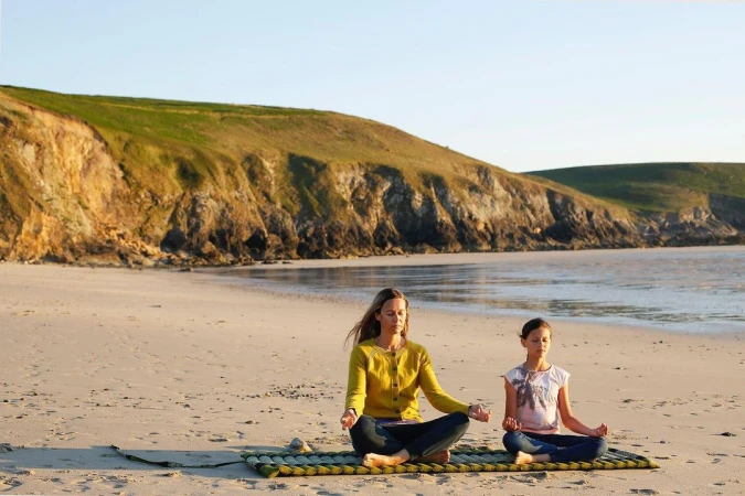 Woman and child practice yoga on beach, sitting cross-legged in meditation pose on mat with ocean and grassy cliffs in background.