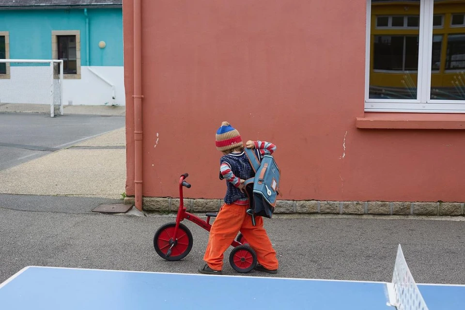 Young child with a backpack and knit hat is near a red tricycle in an urban setting with colorful buildings and a ping pong table.