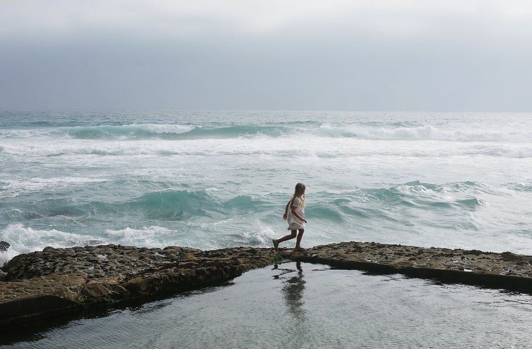 A young girl in a white dress runs along a rocky shoreline with ocean waves, creating a sense of freedom and connection with nature.
