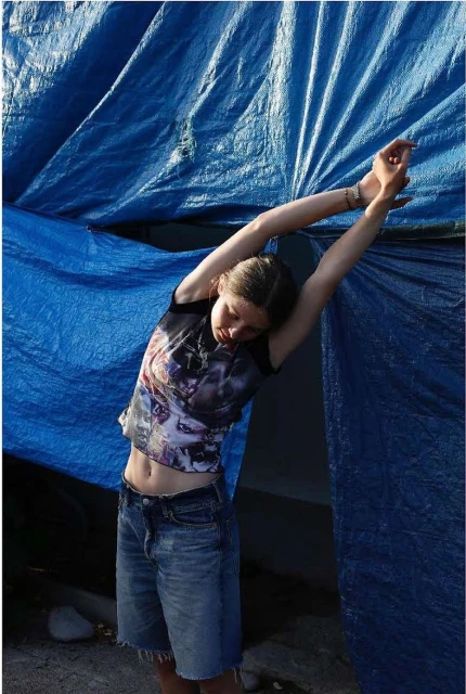 Young woman stretches in front of a blue tarp, wearing a graphic tee and denim shorts in natural light, simple setting, casual pose.