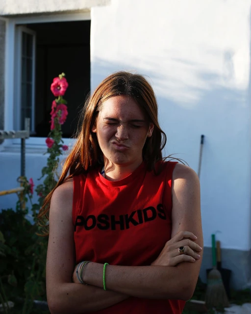 A young woman with crossed arms, wearing a red shirt, stands outdoors with a scrunched face, beside pink hollyhock flowers and a white building.