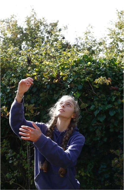 Young woman looking up surrounded by foliage in an outdoor environment. She is wearing a casual outfit with braided hair.