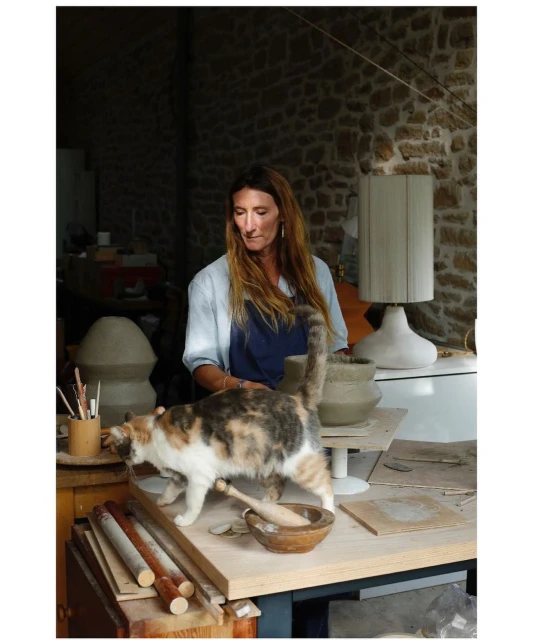 Woman artist working with clay in studio, accompanied by calico cat exploring the worktable in indoor setting, natural light.