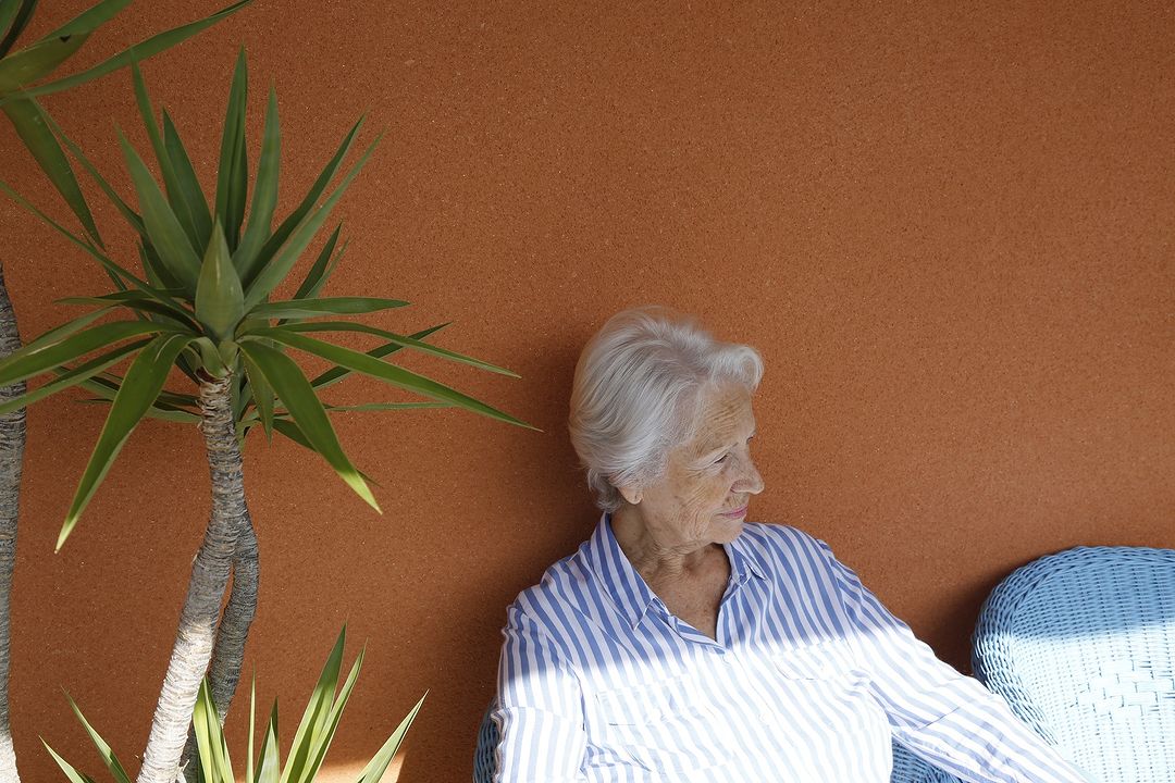 Elderly woman with silver hair sits in outdoor chair next to potted plants against solid orange wall in casual blue and white striped shirt.