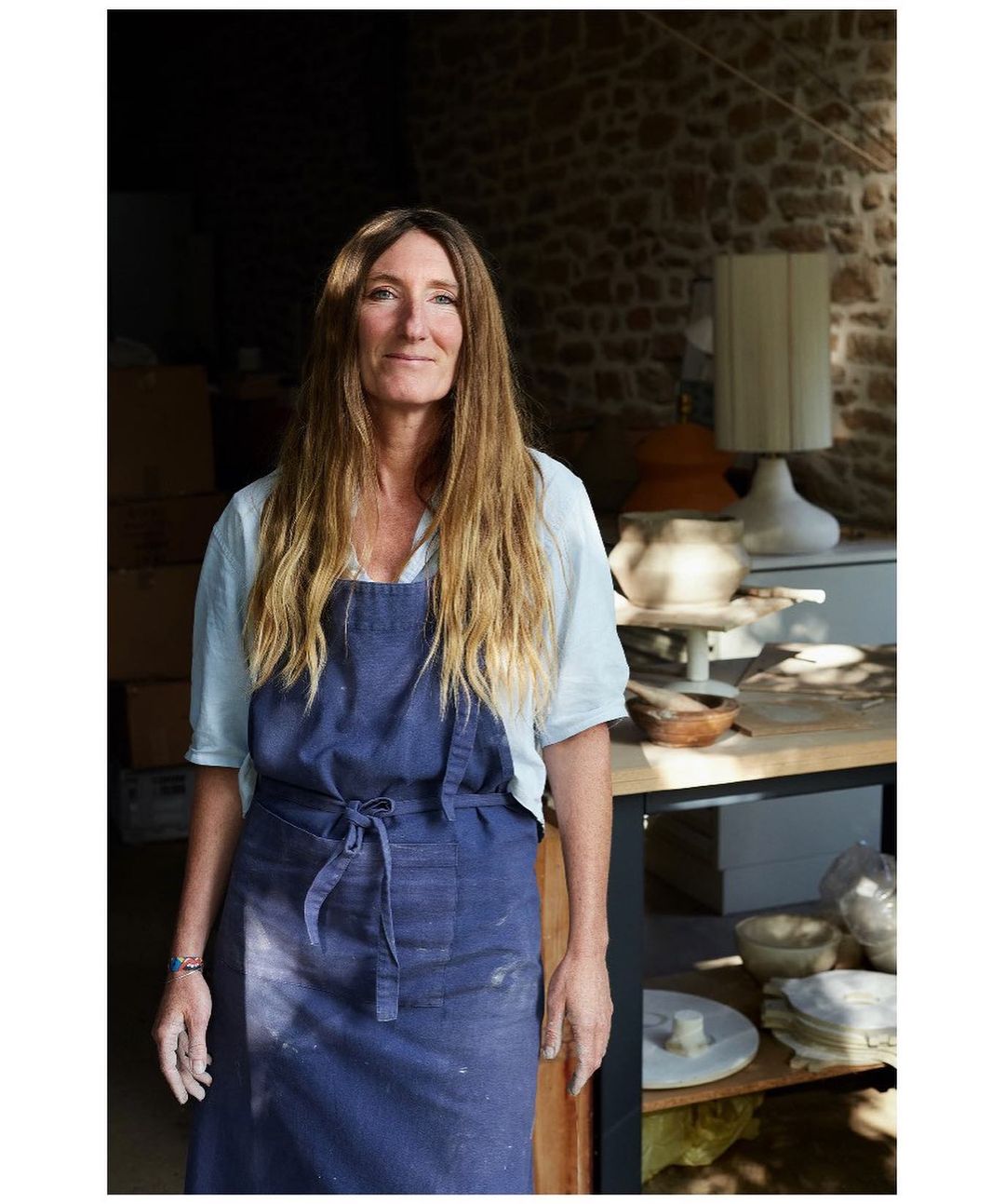 Portrait of a smiling, blonde woman in a blue apron standing indoors in a pottery workshop with pottery items on a wooden table.