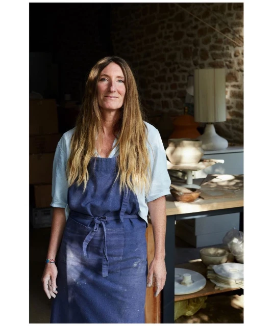 Portrait of a smiling, blonde woman in a blue apron standing indoors in a pottery workshop with pottery items on a wooden table.