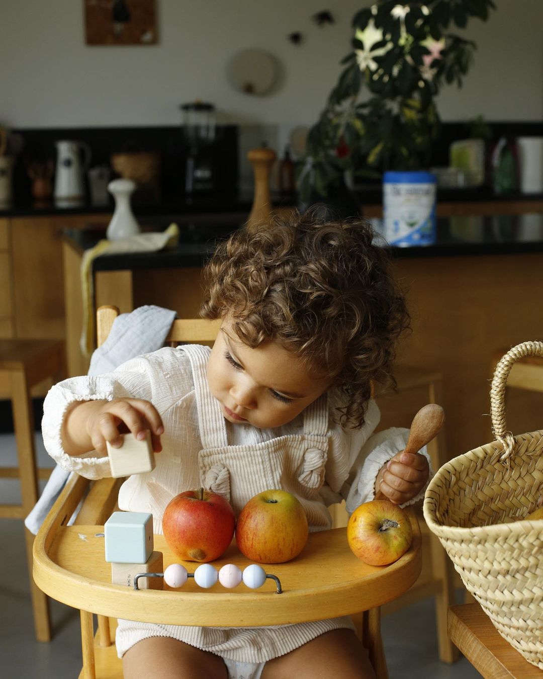 Child sits in high chair playing with wooden blocks, apples, and abacus in a naturally lit home kitchen setting.