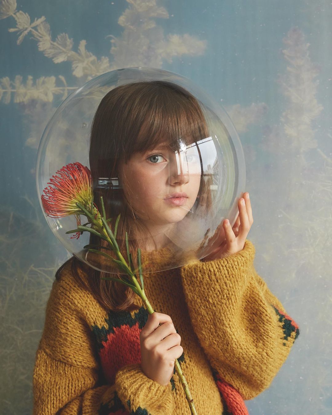 Young girl with brown hair and a clear dome around her head, holding a flower, posing against a blue and white abstract background.