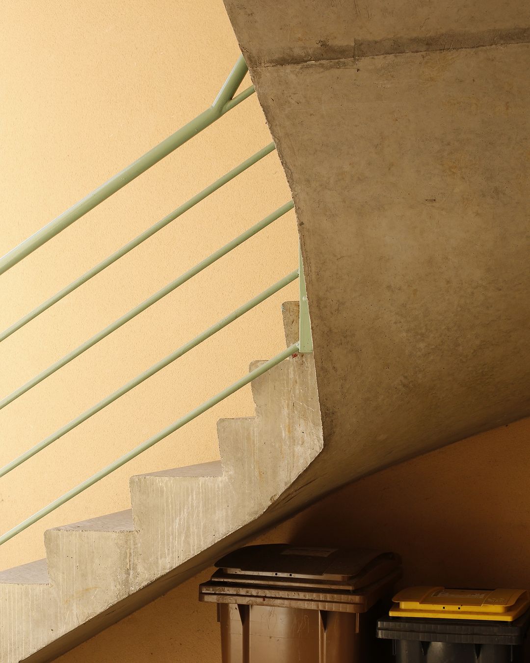 A low-angle shot of a concrete staircase with a green railing, leading up to a corner, with two refuse containers in the foreground.