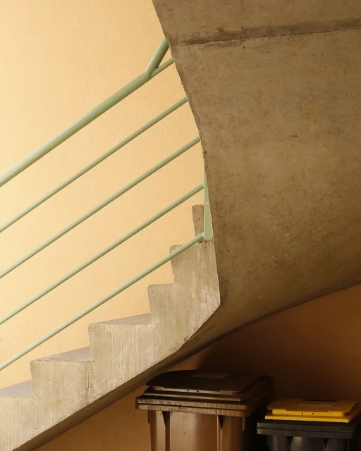 A low-angle shot of a concrete staircase with a green railing, leading up to a corner, with two refuse containers in the foreground.